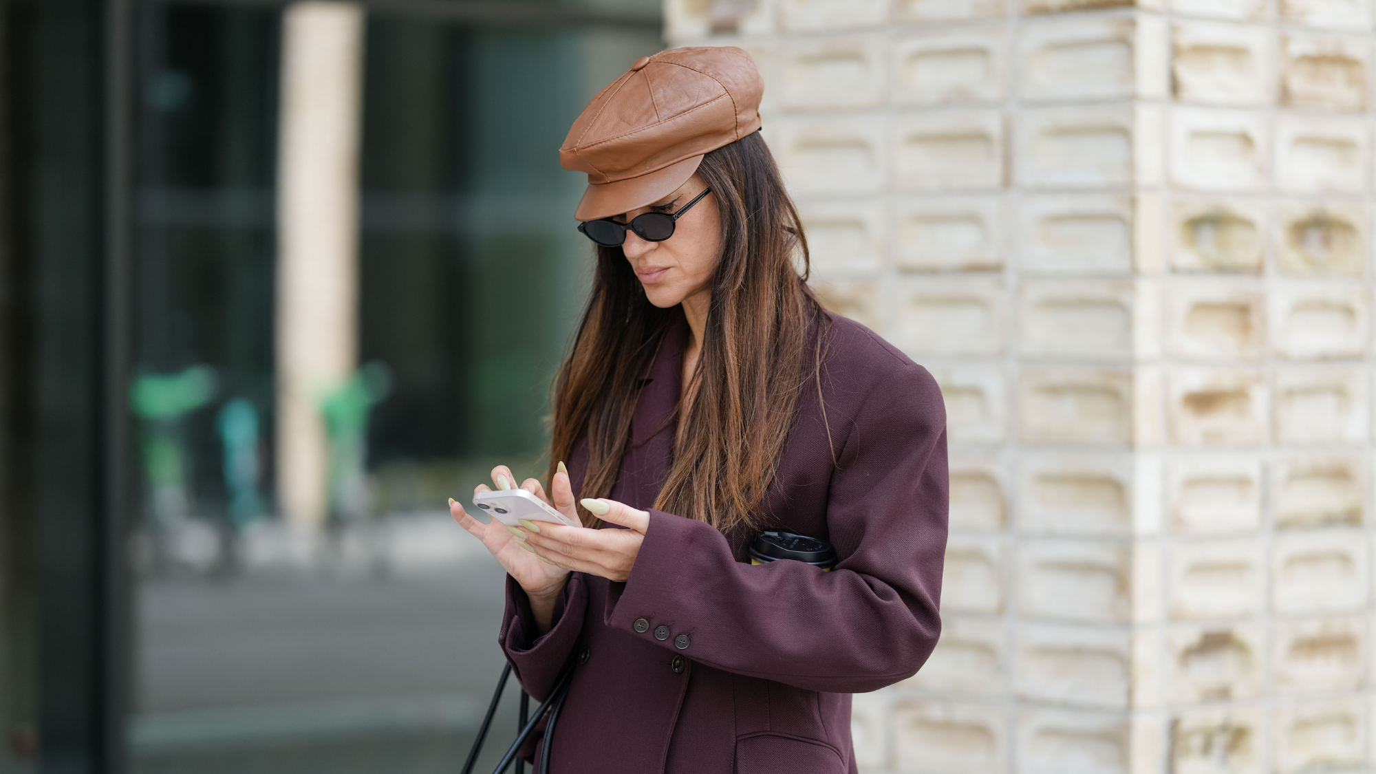 woman wearing a hat, sunglasses and an aubergine-coloured blazer photographed on her phone 