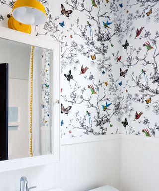 A cottage cor bathroom decorated with butterfly wallpaper and white ship paneling. A white vanity mirror is mounted on the wall with a round yellow wall light installed above