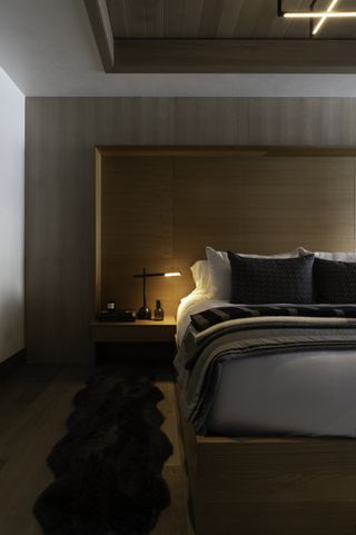 modern bedroom with contemporary wood bed and fitted headboard with storage, wooden floor and wall and section of ceiling, side lamp, white bedlinen with grey bed pillows and blankets, black sheepskin rug