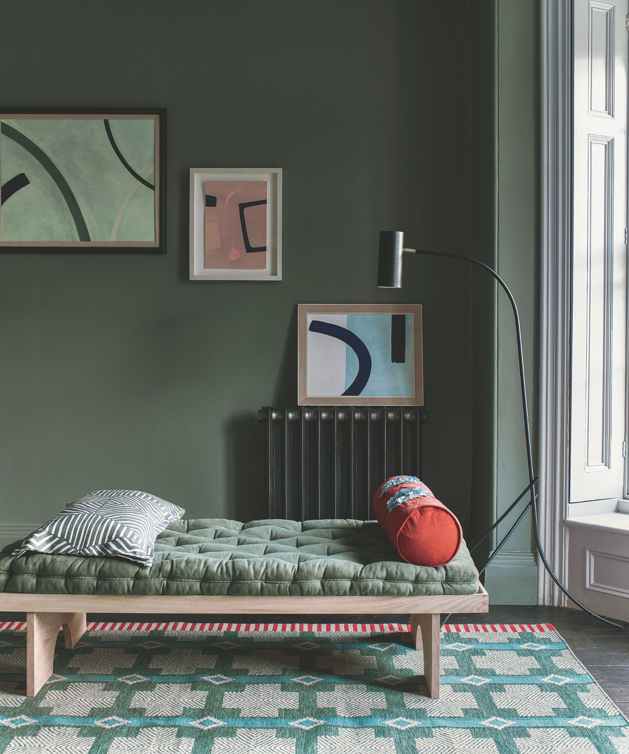 Futon-style bed in a green bedroom with a green mattress, patterned rug, and large window with white shutters to the right. Behind the bed is a radiator with artworks hung above.