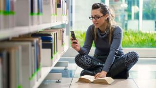 Young woman texts while sitting on library floor next to book shelves.