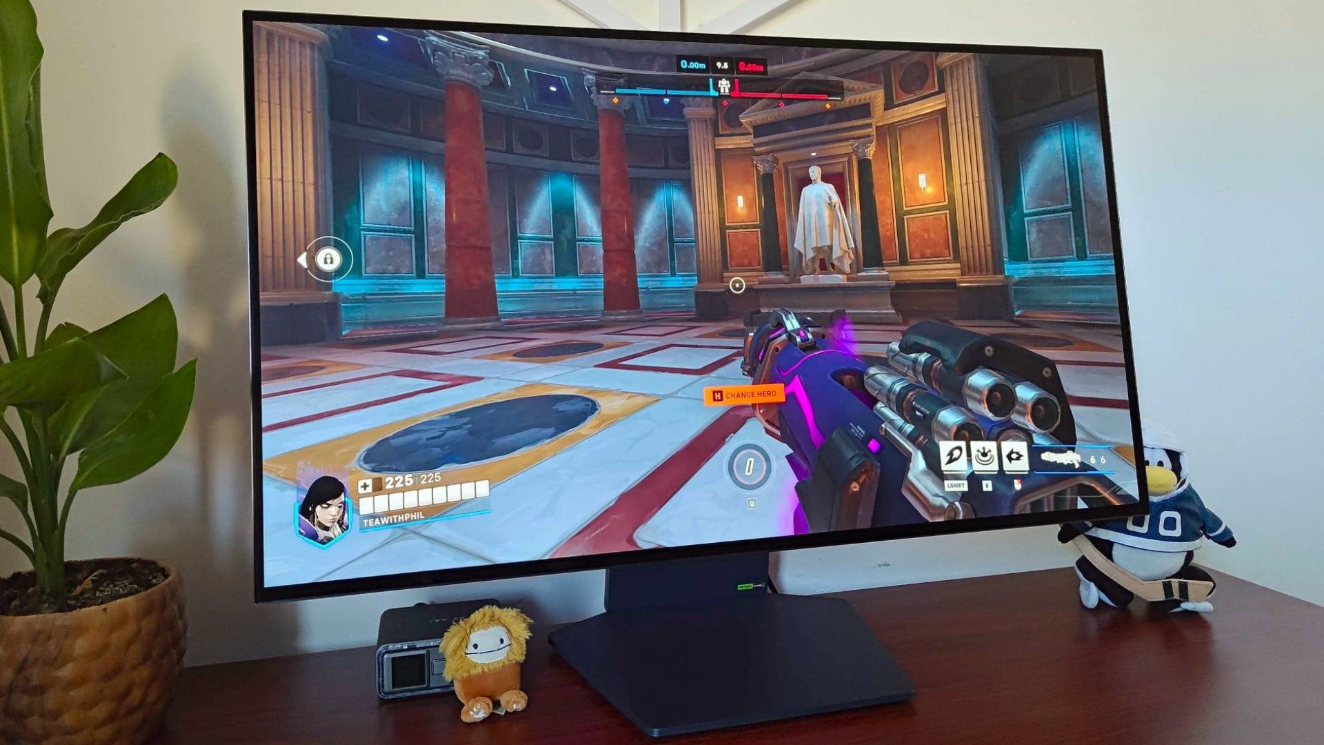 LG UltraGear Dual Resolution monitor on desk next to plant, Bigfoot Squishville, and Club Penguin plush with Overwatch 2 gameplay on screen.