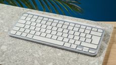 A photograph of the Logitech MX Keys Mini in light gray, positioned on a stone slab with a blue wall in the background.