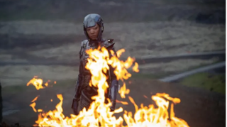 A woman in a black space suit stares into the flames