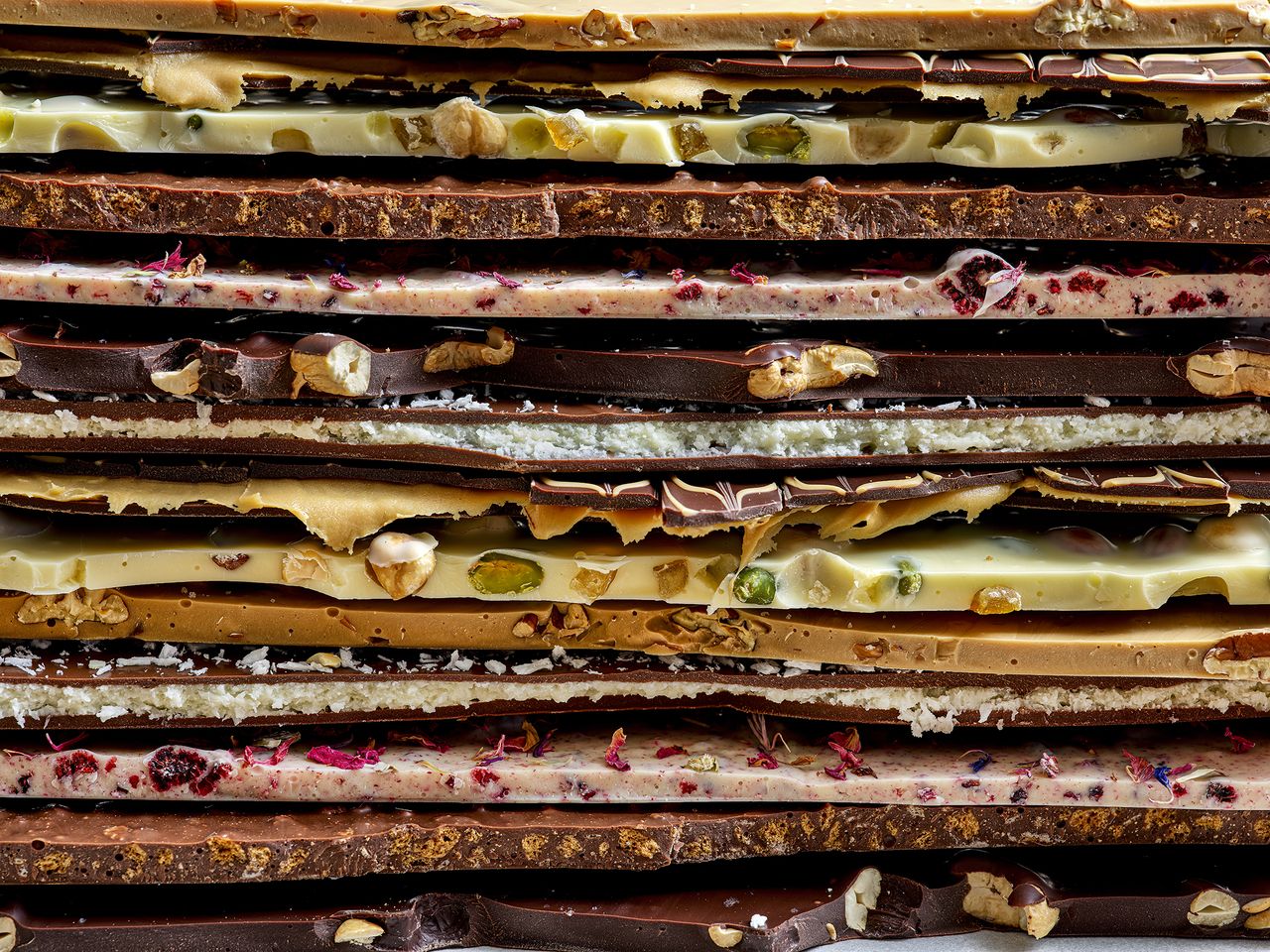 A colorful assortment of layered chocolate bars featuring nuts, fruit, and floral toppings, showcasing various textures and flavors