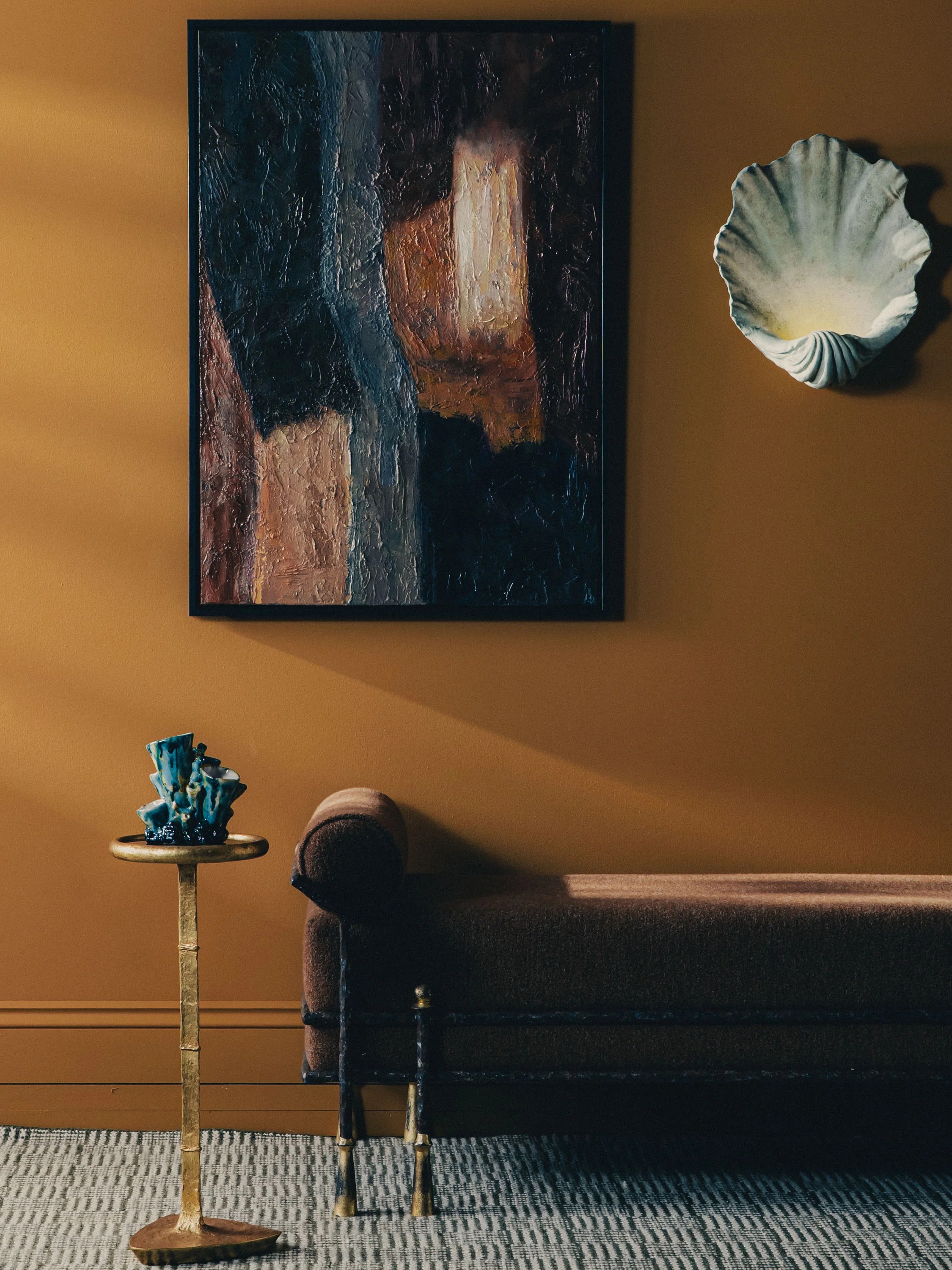 Image of an entryway/hallway painted in a burnt orange color with light gray carpet. There is a brown bench seat, a gold accent table, a shell scone light, and an abstract painting also in the room.