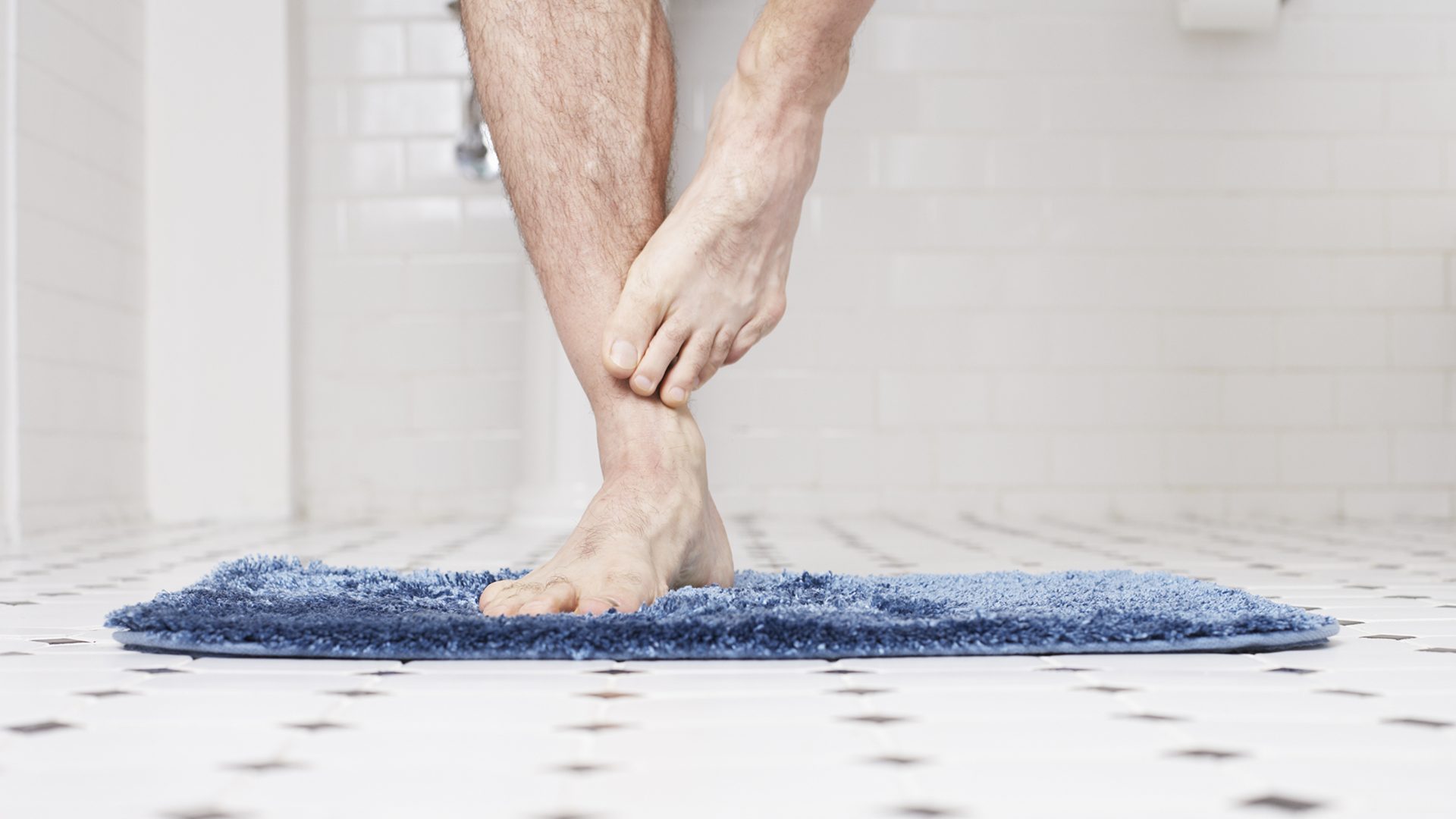 A pair of bare legs standing on a bathmat on a bathroom floor, balancing on one leg.