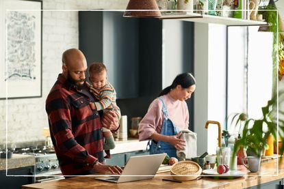 family in kitchen. Father holding baby while looking at laptop while mother is at the sink