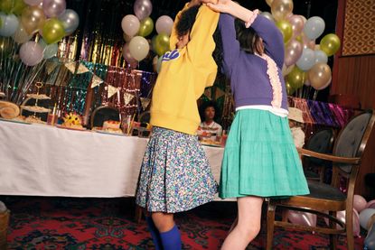 Two girls in party clothes twirl around beside a table laden with party food, their faces obscured 