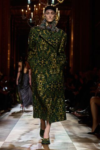 A model wearing an opulent green jacquard coat with a cap and round-toe heels while walking at the F/W 25 Dries Van Noten show during Paris Fashion Week.