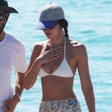 Dakota Johnson stands next to a man wearing a baseball cap, white bikini, floral skirt, and mesh flats.
