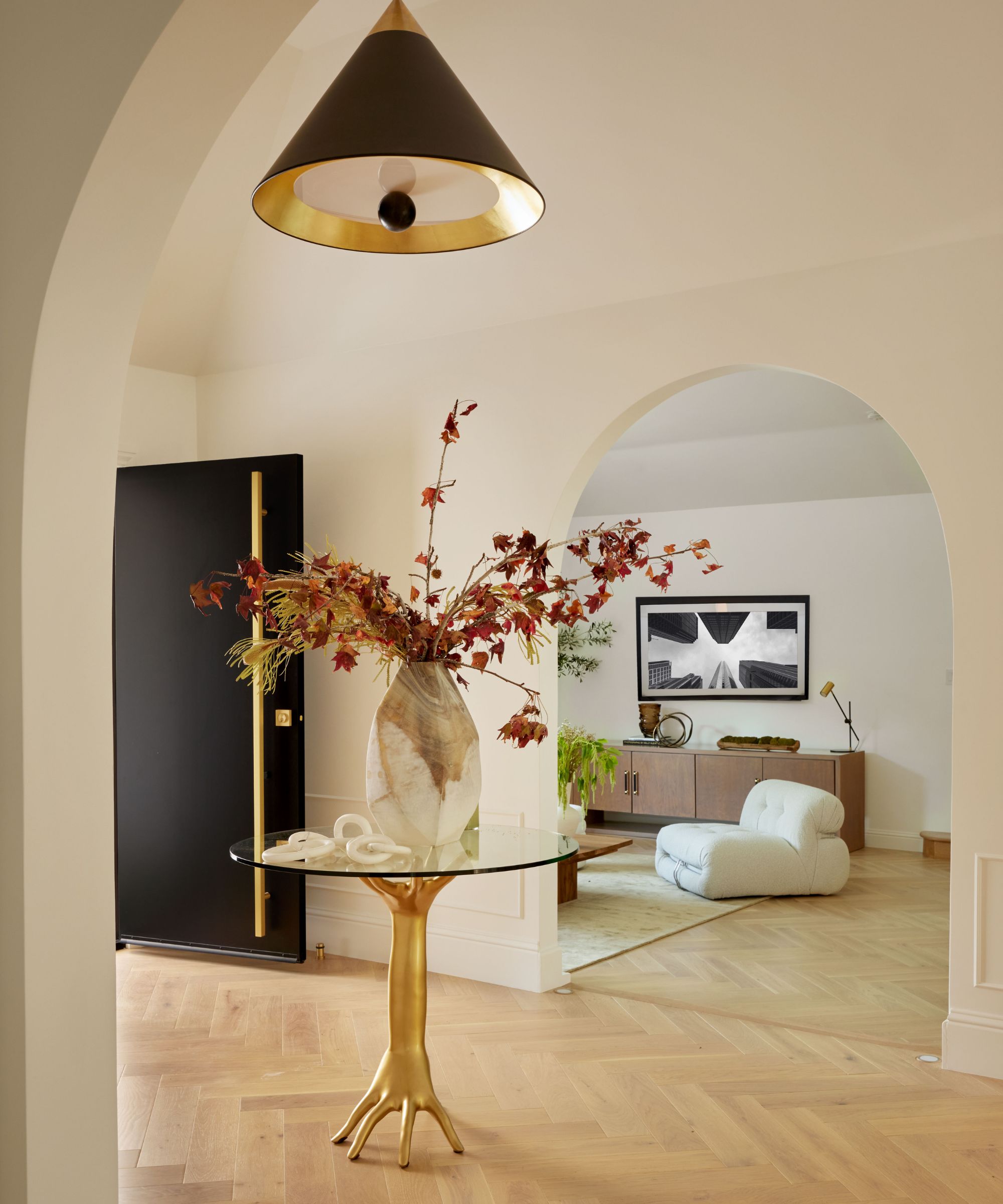 Bright cream entryway with light wooden flooring, round glass table with gold stand, large vase of foliage on top of table, living room in background through arch doorway