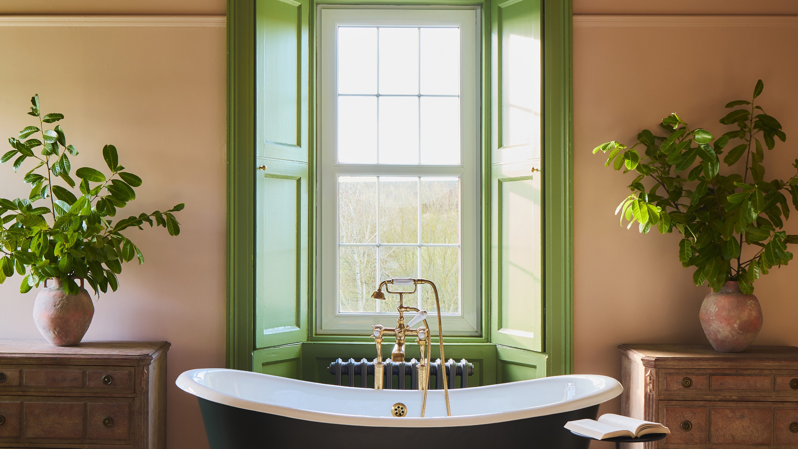 A bedroom colour drenched in Farrow &amp; Ball's Setting Plaster pink paint with a bathtub in the middle of the room and the bay window painted in Farrow &amp; Ball Yeabridge Green