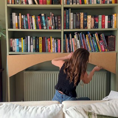 Woman adding plywood arch to bottom of green bookshelf above radiator