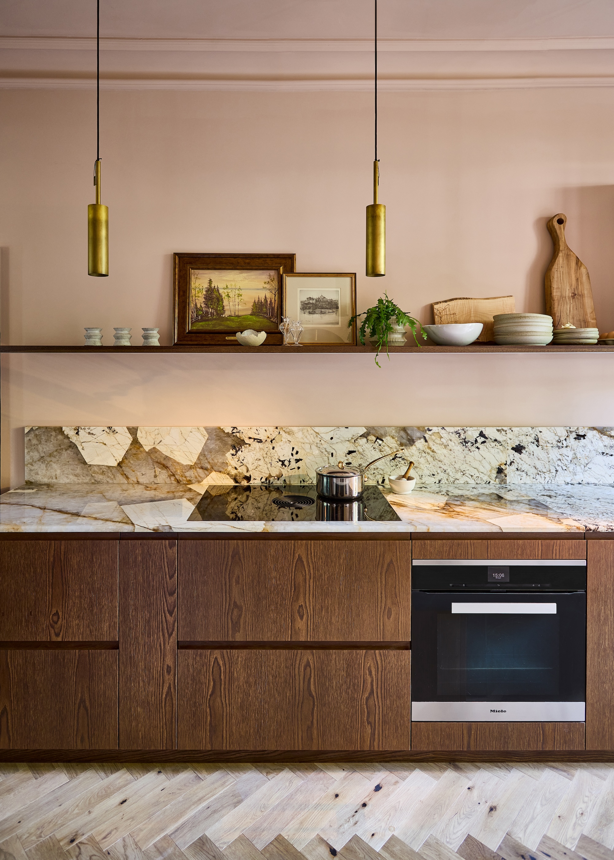 A kitchen with a built in oven as well as open shelving about the kitchen oven and stove. On the shelf is framed artwork as well as bowls and dishes.
