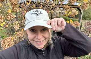 Amy Draiss smiles at the camera, tipping a baseball cap that says "Bee Cups for Thirsty Pollinators"