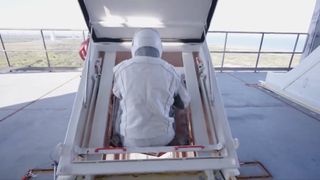 a person wearing a white flight suit gets into a small compartment at the top of a launch tower