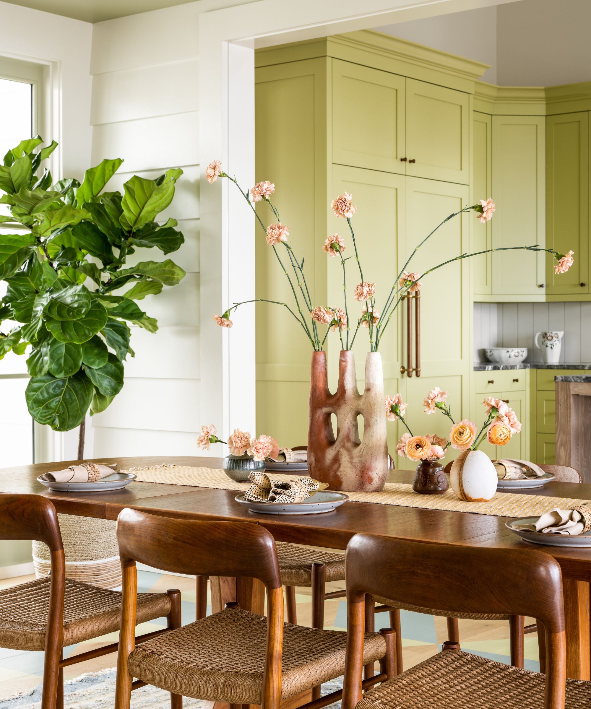 A bright dining room featuring a wooden table with woven chairs and a sculptural clay vase with pink flowers. In the background, a kitchen is visible with light pistachio-green cabinetry and a matching ceiling.