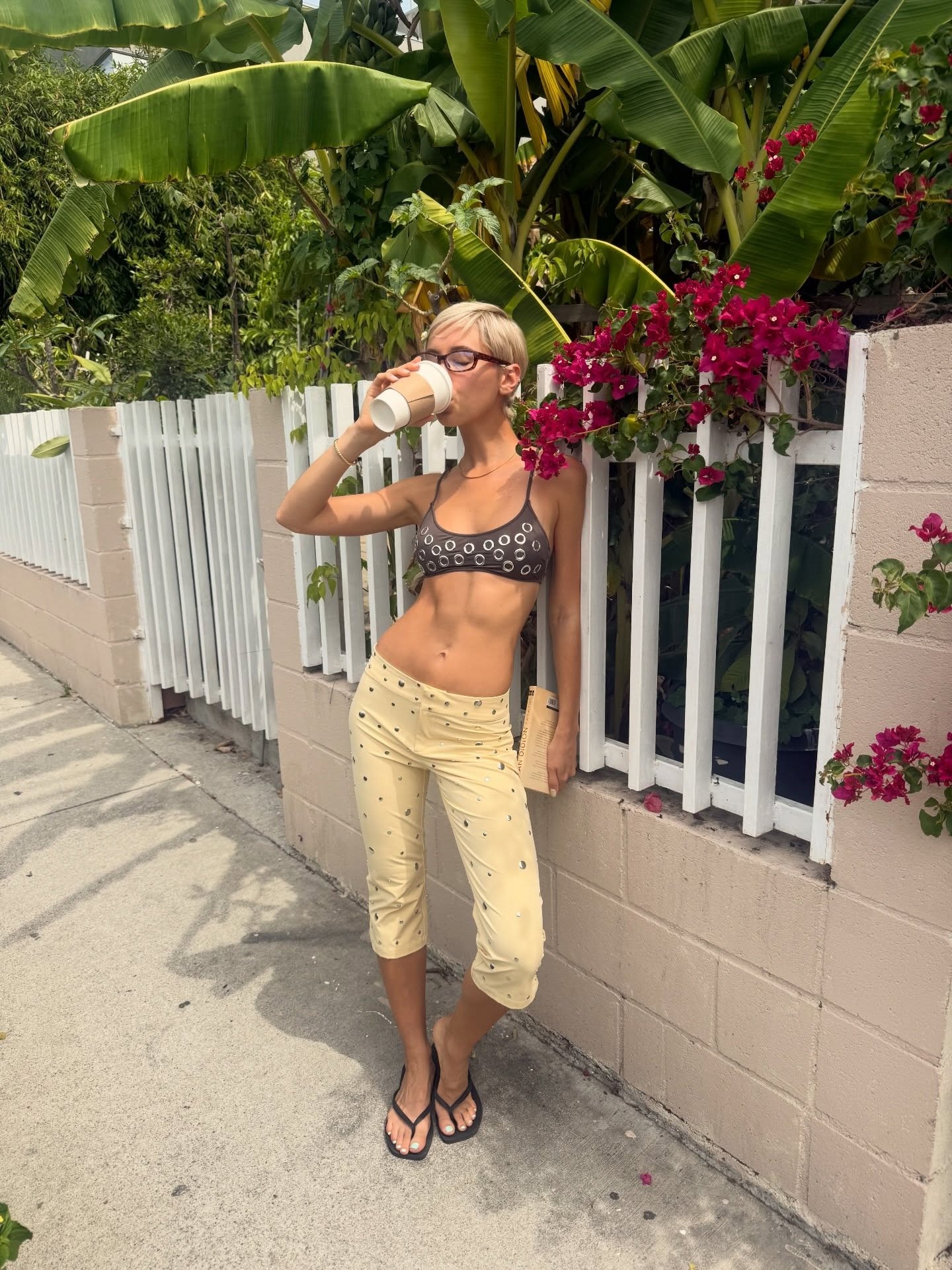 Iris Law drinks coffee outside wearing yellow capri pants and flip flops.