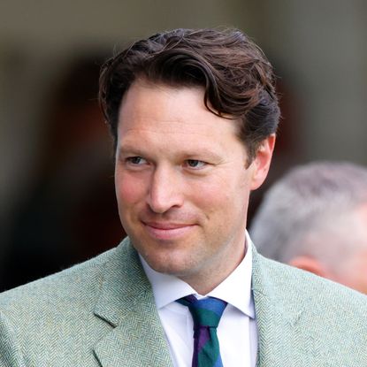 King Charles' equerry Jonny Thompson smiles while wearing a light green suit, a white shirt, and a striped tie