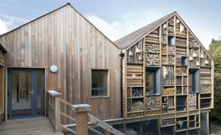 Mellor Primary School, daytime image, wooden barn style building, grey framed entrance door and windows, wooden framed handrail leading down to the ground level, one half of the building has wooden log design in sections and bird houses attached, wooden floor/platform to the balcony, black drain pipe, trees to the right hand side, gravel path, wire fence front to the lower level, cloudy blue sky