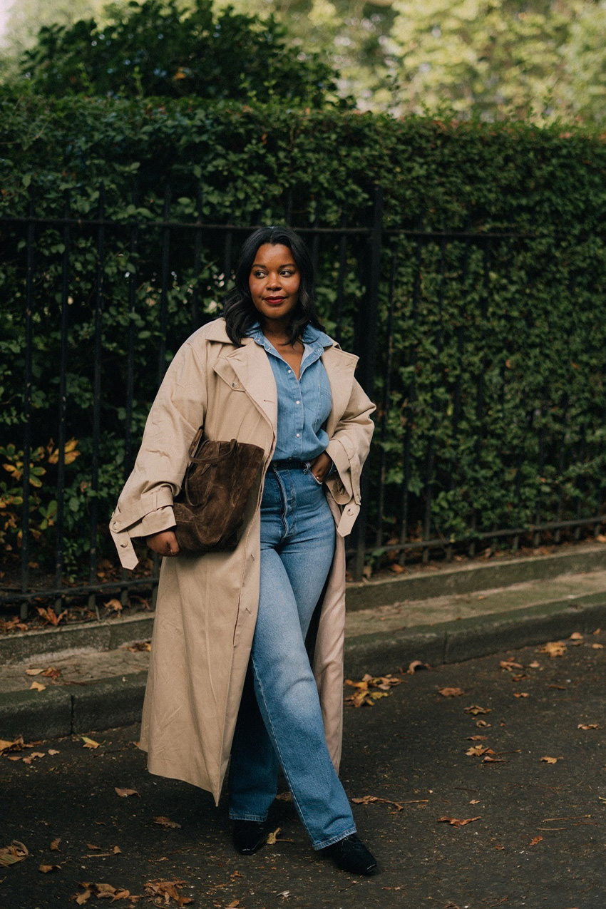 How to wear skinny jeans is shown in a photo of a woman standing on the sidewalk wearing a tan trench coat over a denim button-down shirt with matching skinny jeans, black ankle boots, and a brown suede handbag
