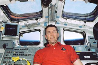 Astronaut Rick D. Husband wears a red t-shirt. Behind him are various control panels and several large windows where you can see Earth in the background.