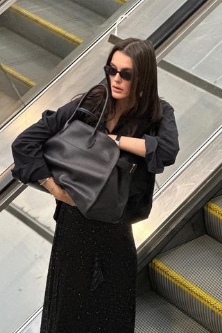 How to make a button-down shirt look more expensive is shown in a photo of a woman standing on an escalator wearing black sunglasses with a black oversized button-down shirt, a black watch, a black tote bag, a black maxi skirt, and black flip flops