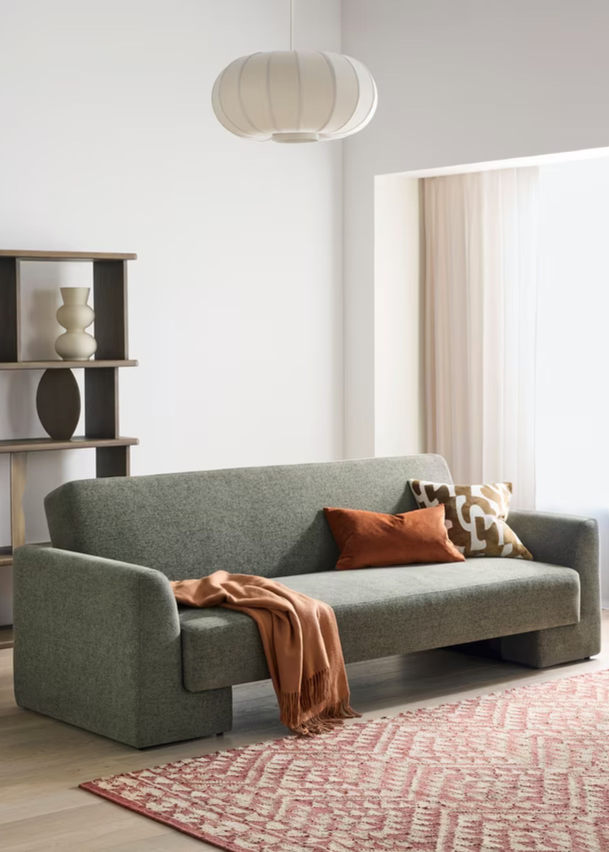 A green 3-seater sofa bed styled in a white-painted living room with an orange cushion and throw on the sofa with another patterned cushion in front of a patterned rug, with a rice paper pendant above