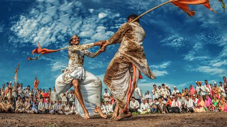 Two men in traditional attire engage in a vigorous dance with flags, surrounded by an audience in colorful clothing under a vibrant sky