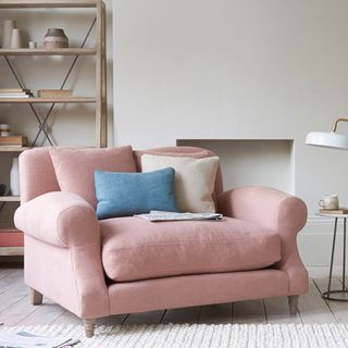 a pink loveseat sofa with pink, cream and blue cusions, in a living room with an open wooden display unit against the wall