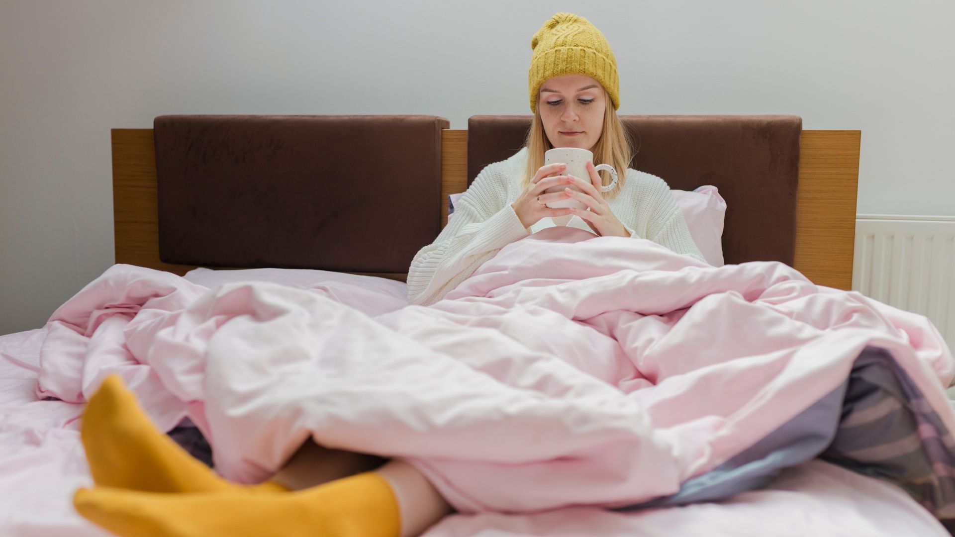 A woman wearing a beanie hat and socks sis under a duvet in bed while drinking from a mug.