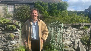 Monty wearing a cream coat and standing in front of a picture-perfect country cottage and garden