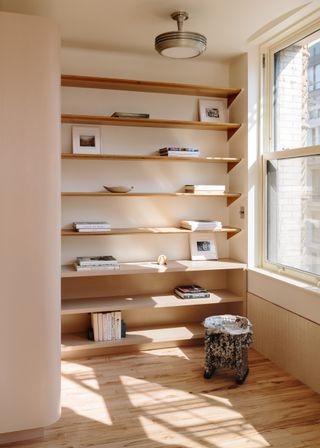 A wall with floor to ceiling wooden shelving, a round stool and a window.