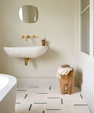 Neutral, calming bathroom space with cream color palette, textured sink, irregular rounded mirror, wooden stool, patterned cream and black tiles, cream painted walls.