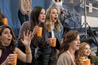 A group of women (Beth Dover as Talia, Jackie Tohn as Esther, Kristen Bell as Joanne, Elise Greene as Leah, Rachel Rosenbloom as Rachel) look surprised while holding orange cups and sitting in the stands in a gym, in episode 107 of Nobody Wants This.