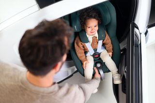 A smiling toddler sits in a Maxi Cosi car seat while a parent slides it to face the car door