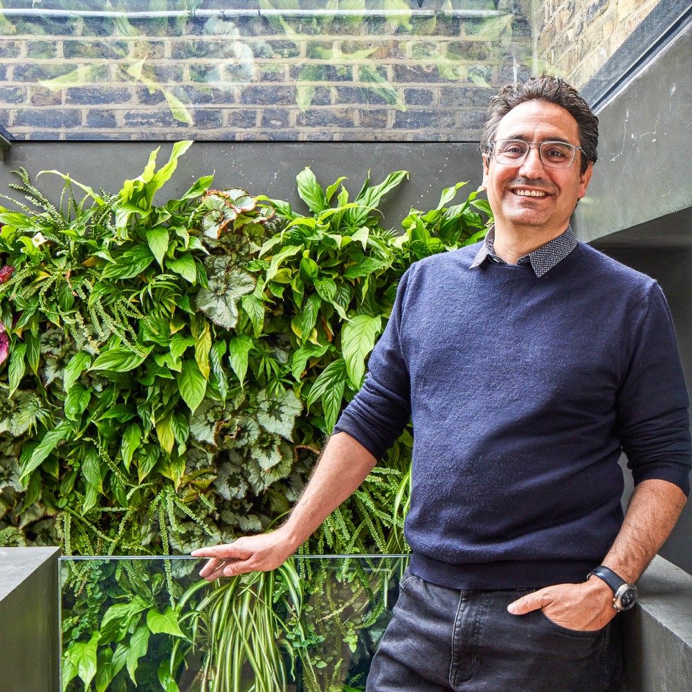 male with short dark hair, glasses, wearing navy jumper, shirt and jeans stood in building with glass roof and green living wall