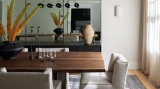 Image of a wooden dining table with off-white fabric chairs around it and a large mirror on the back wall. There is a white sconce light next to the mirror.