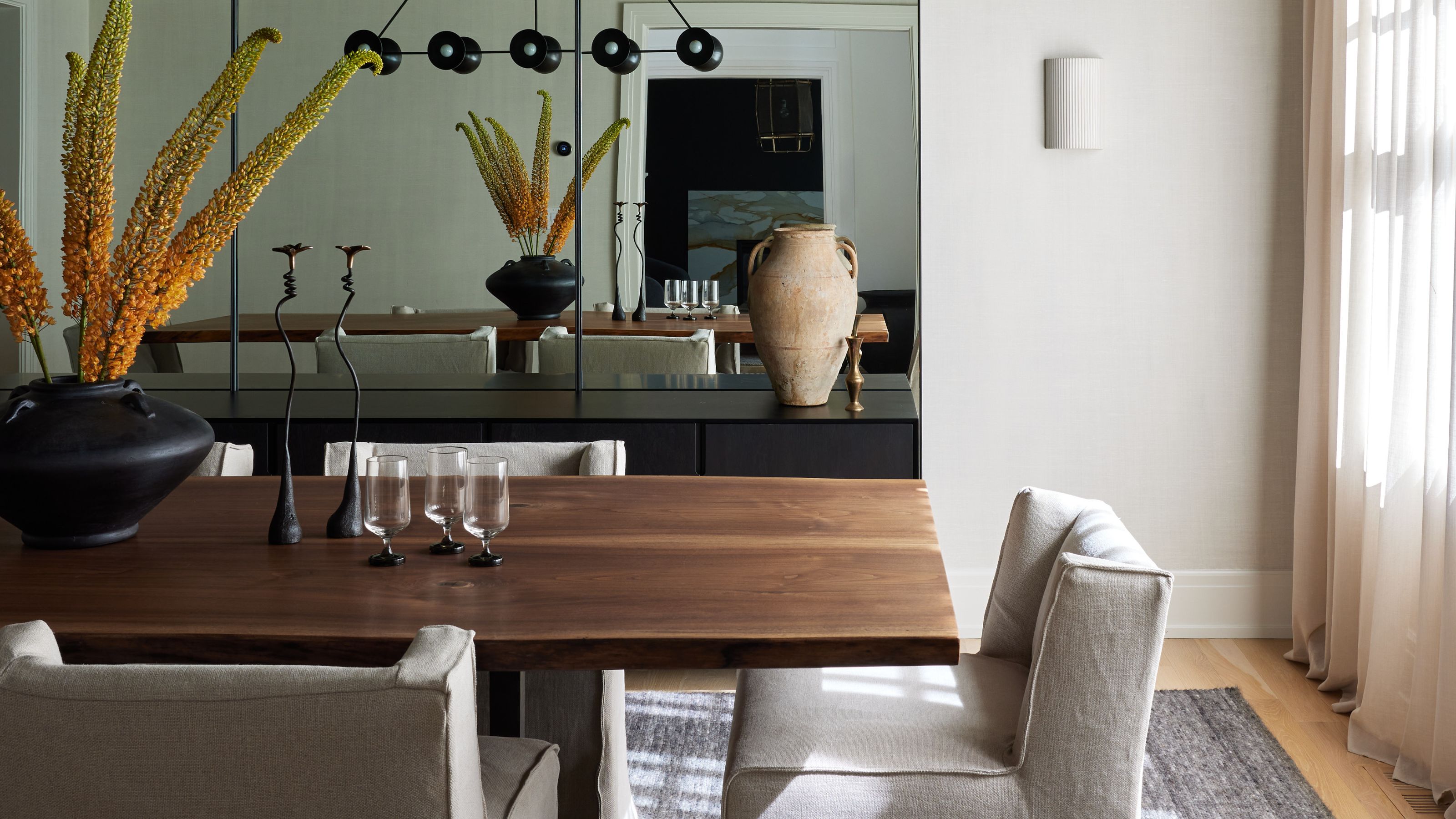 Image of a wooden dining table with off-white fabric chairs around it and a large mirror on the back wall. There is a white sconce light next to the mirror.
