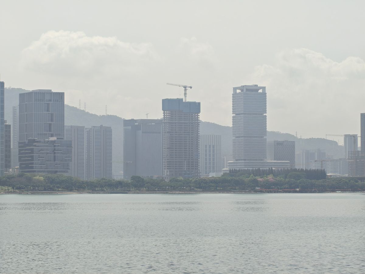 A telephoto shot of a city skyline across a body of water, featuring several modern skyscrapers in various stages of completion. One prominent building in the center is under construction with a yellow crane on top. The background shows a hazy green mountain range under a cloudy, bright sky.