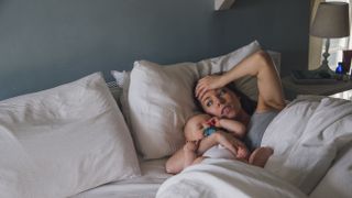 A woman with dark hair lies in bed with her baby while feeling sleep deprived