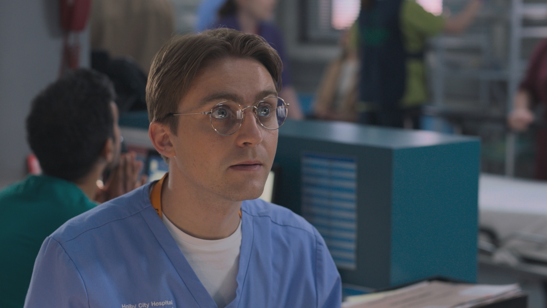 Cam sitting at Holby ED's reception area, wearing glasses and staring into space.