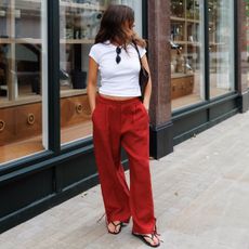 British style influencer Kim Turkington poses on a London sidewalk wearing a white T-shirt, brown woven bag, red linen pants, and flip-flop sandals