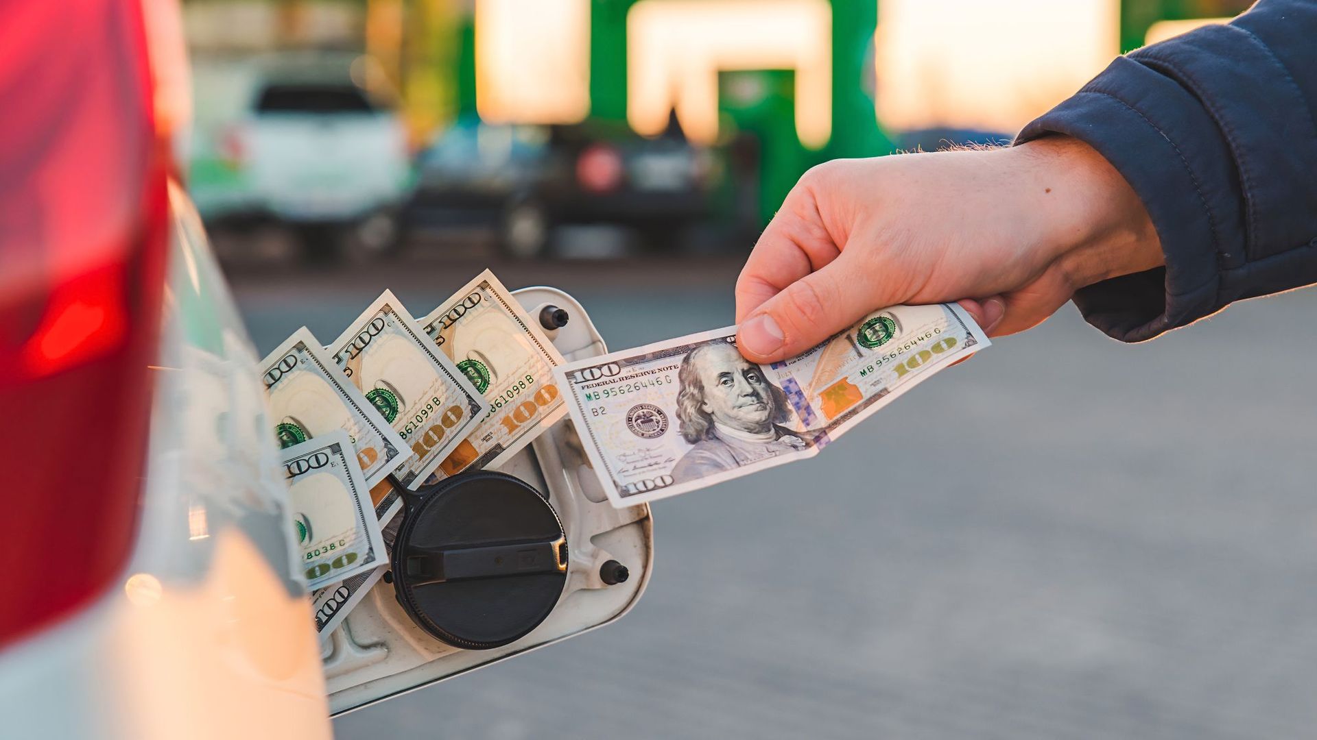 A hand putting $100 bills in a car's gas tank.