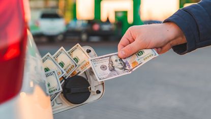 A hand putting $100 bills in a car's gas tank.