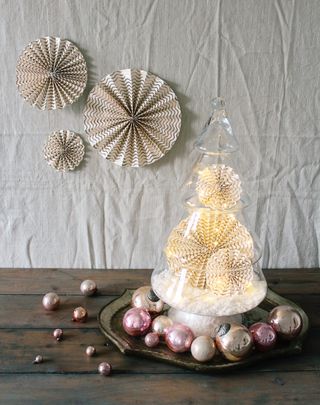glass Christmas table top tree with faux snow and paper decorations, baubles around it on a table