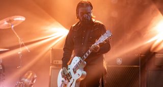 Billy Duffy of the Cult wears a black western shirt and plays his signature Gretsch White Falcon onstage in london, with orange stage lights in the background.