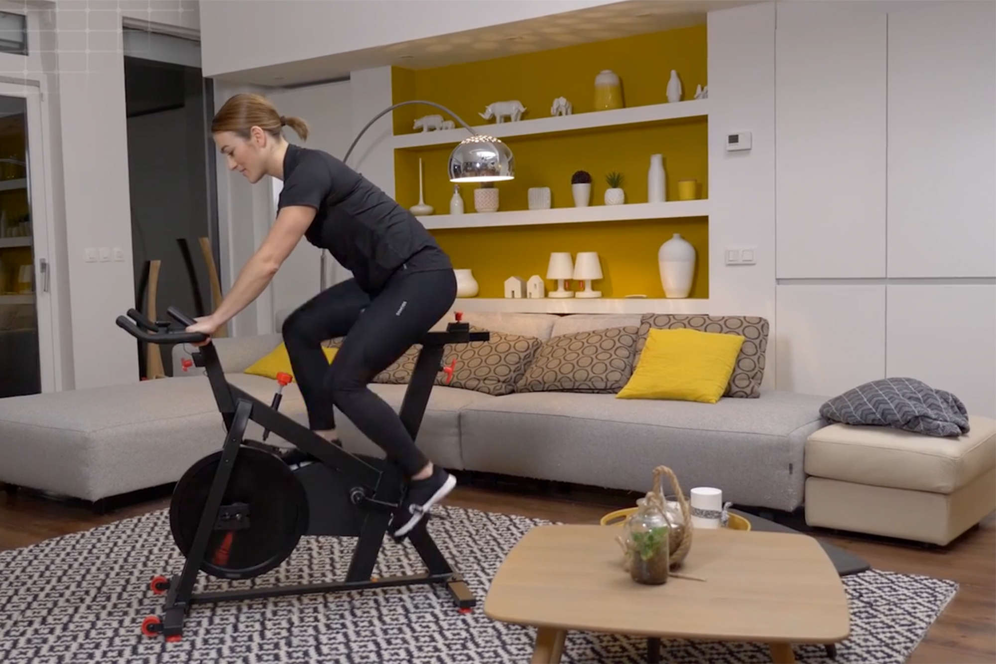 female riding the Domyos basic exercise bike 100 in a living room setting, which is decorated in a mustard yellow and cream with a grey sofa just behind.