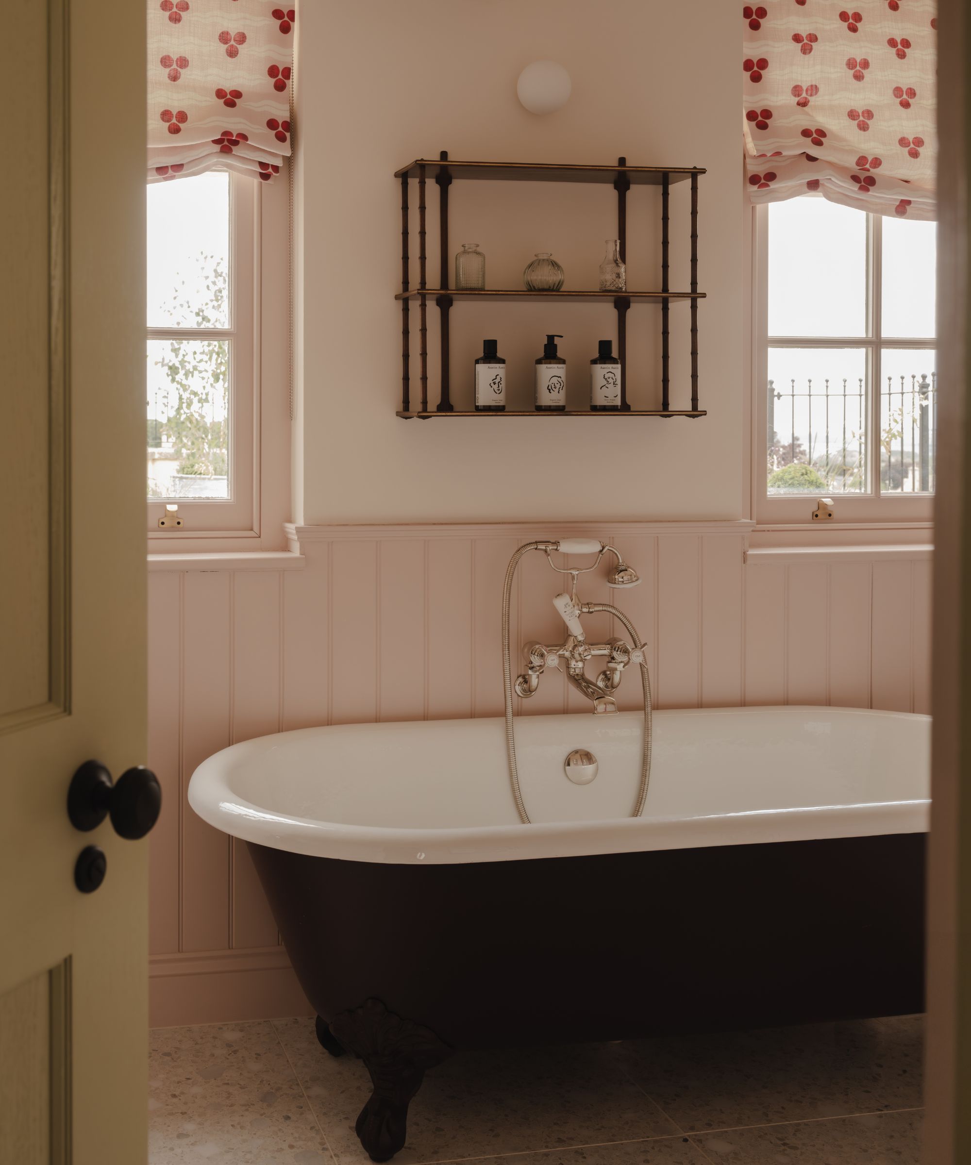 a pale pink bathroom with tongue and groove paneling, two floral window blinds, a cast iron roll top bath and antique wall shelf